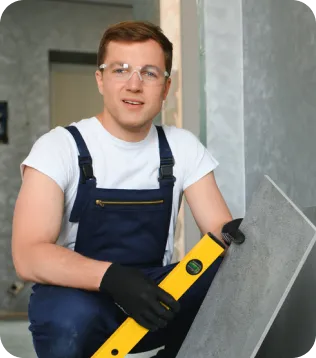 Junger Fliesenleger in Latzhose mit Wasserwaage und Fliese auf der Baustelle