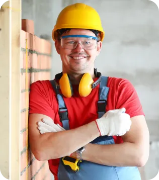Lächelnder Maurer und Betonbauer mit gelbem Schutzhelm, Schutzbrille und Gehörschutz vor einer Ziegelwand auf der Baustelle