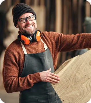 Tischler mit Mütze, Gehörschutz und Schürze begutachtet ein Holzbrett in der Schreinerei