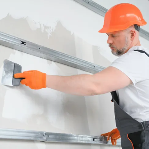 Ein Trockenbauer mit orangem Bauhelm und Handschuhen verspachtelt eine Wand, an der Metallprofile zur Befestigung angebracht sind. Er trägt Arbeitskleidung und arbeitet konzentriert an der glatten Oberfläche.