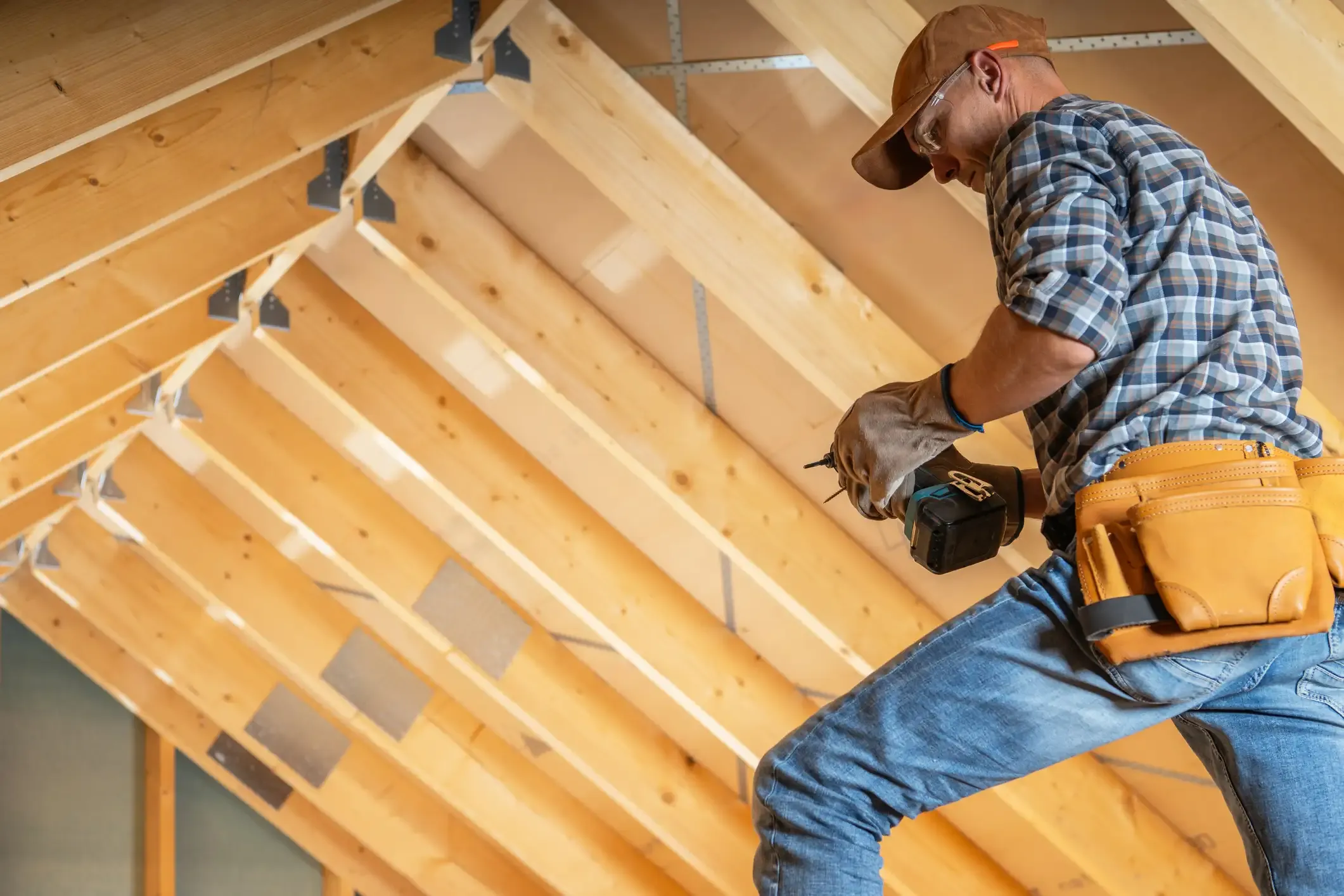 Handwerker mit Werkzeuggürtel und Akkuschrauber beim Dachausbau in einem unfertigen Dachgeschoss. Im Hintergrund ist die offene Dachkonstruktion aus neuen Holzsparren und die Unterdeckbahn der Dachschräge sichtbar.