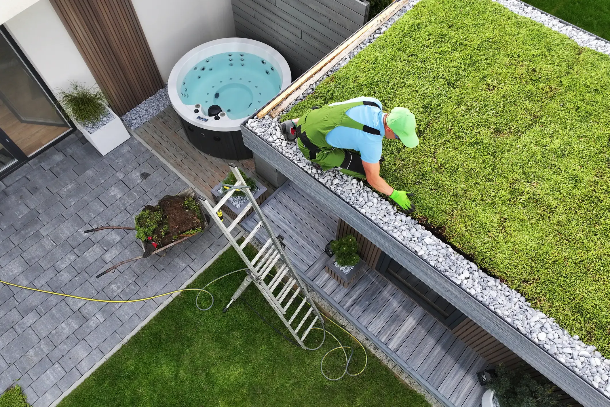 Vogelperspektive auf einen Handwerker in grüner Arbeitskleidung, der eine extensive Dachbegrünung mit Sedum-Matten auf einem modernen Flachdach anlegt. Am Dachrand verläuft ein Kiesstreifen aus hellen Kieselsteinen, während im Garten darunter eine Leiter, eine Schubkarre und ein Whirlpool zu sehen sind.