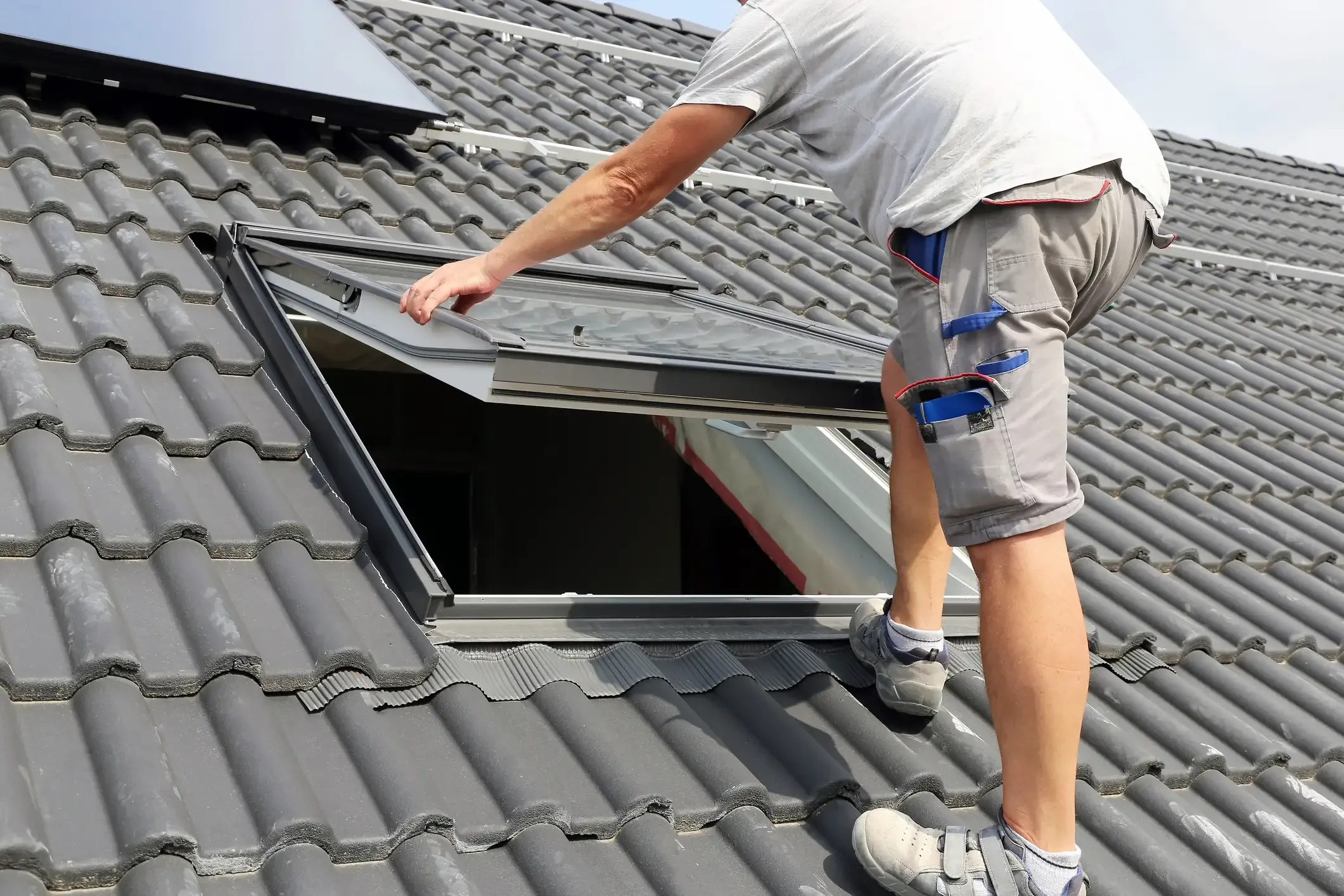 Ein Dachdecker führt einen Dachfenstereinbau auf einem mit dunklen Pfannen gedeckten Schrägdach durch. Er setzt den beweglichen Fensterflügel in den bereits montierten Einbaurahmen des neuen Dachfensters ein, während im oberen Bereich Photovoltaikmodule sichtbar sind.