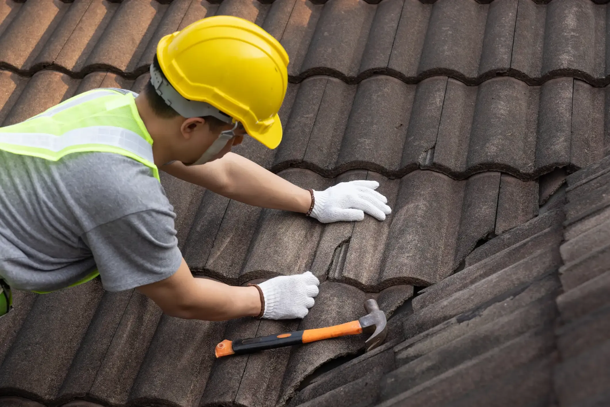 Dachdecker mit gelbem Schutzhelm und Arbeitshandschuhen führt eine Dachreparatur auf einem Ziegeldach durch. Er begutachtet eine beschädigte, gerissene Beton-Dachpfanne, während neben seinen Händen ein Zimmermannshammer auf den dunklen Dachziegeln bereitliegt.