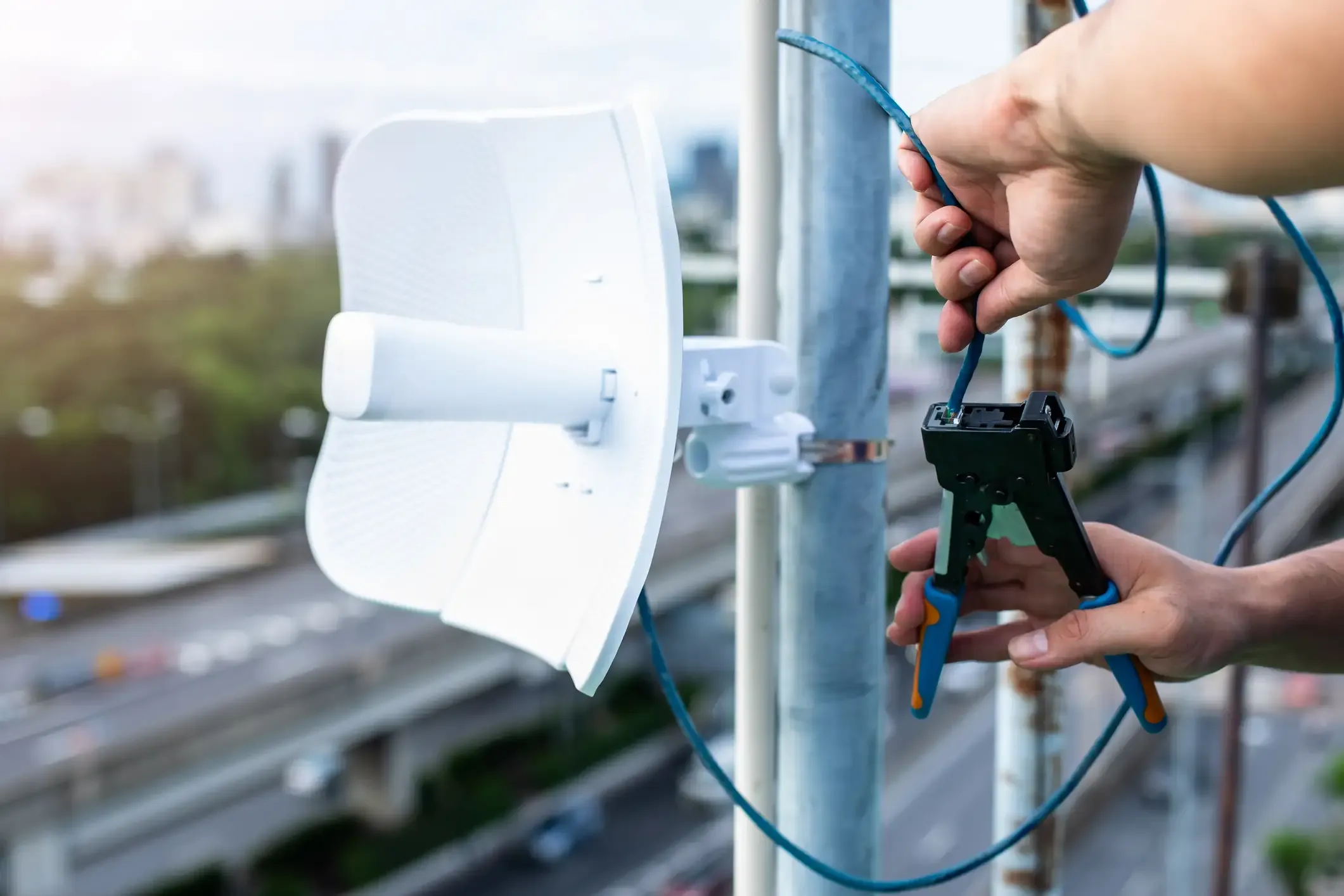 Ein Kommunikationstechniker installiert ein blaues Netzwerk- beziehungsweise Ethernet-Kabel mit einer Crimpzange an einem Metallmast. Direkt daneben ist eine weiße Parabol-Richtfunkantenne für moderne Kommunikationstechnik im Außenbereich montiert.