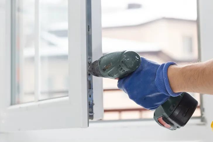 Ein Fensterbauer mit blauen Handschuhen nutzt einen Akkuschrauber, um bei einer Fensterreparatur einen Beschlag am Fensterrahmen zu befestigen.