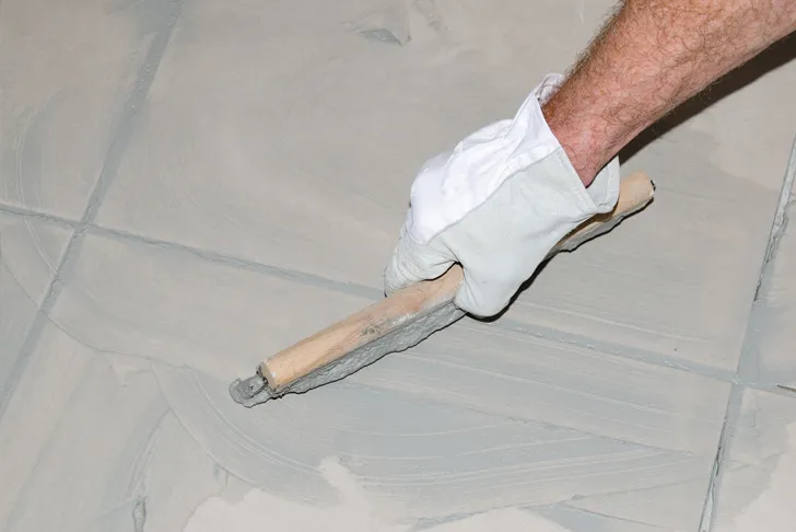 Verfuger trägt Fugenmasse mit einem Fugbrett auf frisch verlegte Fliesen auf. Die Hand ist mit einem weißen Arbeitshandschuh geschützt.