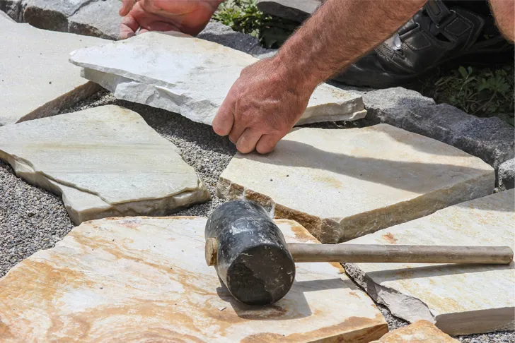 Natursteinleger verlegt Natursteinplatten für einen Gehweg oder eine Terrasse. Ein Gummihammer liegt auf den Steinen, während er die Platten sorgfältig anpasst. Der Untergrund besteht aus verdichtetem Kies.