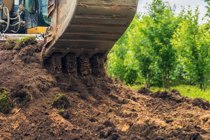 Ein Bagger hebt bei Baggerarbeiten in einem Garten Erde vom Boden aus. Die Schaufel des Baggers ist mit dunkler, feuchter Erde gefüllt. Im Hintergrund sind junge, grüne Bäume sichtbar.