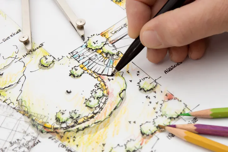 Eine Nahaufnahme einer Hand, die mit einem schwarzen Stift an einem farbigen Landschaftsplan arbeitet. Auf dem Plan sind Details wie Bäume, Wege und Wasserflächen in bunten Farben dargestellt. Die Szene umfasst auch Zeichenwerkzeuge wie Zirkel und Bleistifte, die auf dem Papier liegen, was auf präzise Planungsarbeit im Bereich der Gartengestaltung hindeutet.