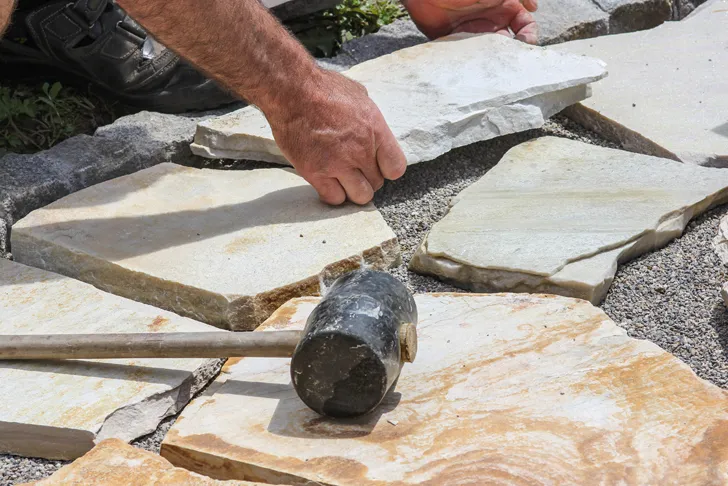 Ein Gartenbauer verlegt Natursteinplatten auf einem Untergrund aus Kies, um eine Terrasse oder einen Gehweg zu gestalten. Die Hände des Gartenbauers positionieren die Platten sorgfältig, während ein Gummihammer daneben liegt. Das Bild zeigt den handwerklichen Prozess von Pflasterarbeiten bei der Gartengestaltung.