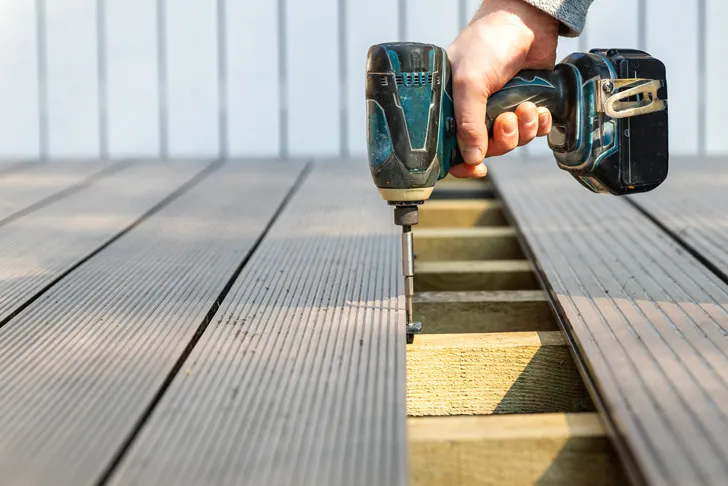 Die Hand eines Terrassenbauers hält einen Akkuschrauber und befestigt eine Terrassendiele auf einem Holzunterbau. Die Terrasse besteht aus langlebigem, wetterfestem Material. Im Hintergrund ist eine weiße Wand sichtbar. Das Bild zeigt den Bauprozess einer Holzterrasse, inklusive Verwendung von Werkzeugen und Baumaterialien.