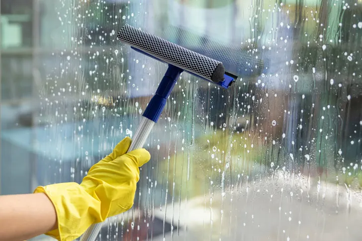 Eine Person mit gelben Gummihandschuhen reinigt bei einer professionellen Glasreinigung eine große Fensterscheibe mit einem Abzieher und entfernt dabei Seifenwasser.