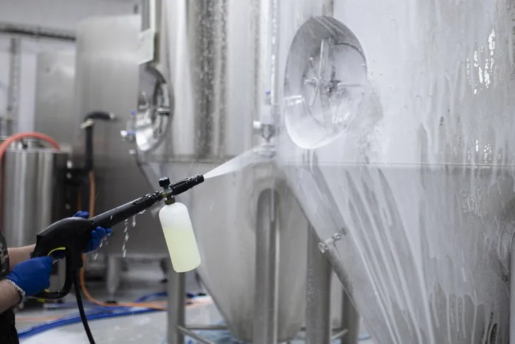 Eine Fachkraft mit blauen Schutzhandschuhen reinigt mit einem professionellen Reinigungssystem die Oberfläche eines großen Edelstahltanks in einer industriellen Produktionshalle.
