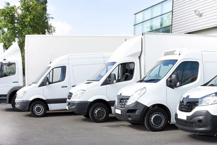 Mehrere weiße Transporter und Lieferfahrzeuge eines Transportunternehmens sind nebeneinander auf einem Parkplatz vor einem modernen Gewerbegebäude abgestellt.