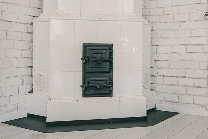 Ein weiß gekachelter Kachelofen in einer Zimmerecke. Der Ofen hat eine schwarze gusseiserne Doppeltür in der Mitte. Er steht vor einer weiß gestrichenen Backsteinwand auf einem hellen Holzboden. Vor dem Ofen liegt eine schwarze Bodenplatte zum Schutz des Bodens.
