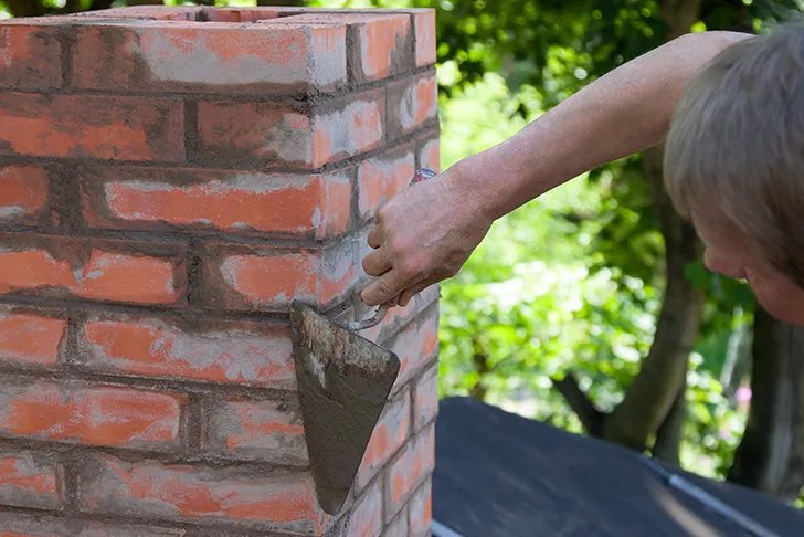 Ein Schornsteinbauer bei der Errichtung eines neuen Schornsteins aus roten Backsteinen. Die Person trägt mit einer Maurerkelle grauen Mörtel auf die Fugen des Mauerwerks auf. Der Schornstein befindet sich im Außenbereich auf einem Dach, im Hintergrund sind grüne Bäume sichtbar.