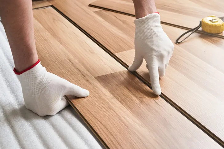 Professionelle Bodenverlegung: Ein Handwerker mit weißen Handschuhen passt Laminatdielen auf einem Boden an. Der Fokus liegt auf der genauen Ausrichtung und Verlegung der Dielen.