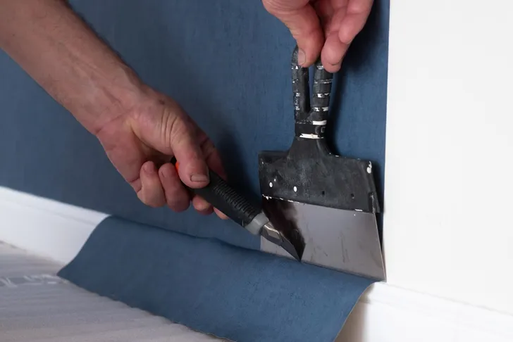 Nahaufnahme von Tapezierarbeiten: Ein Handwerker schneidet mit einem Cuttermesser und einem Tapetenglätter eine blaue Tapete an einer Fußleiste.