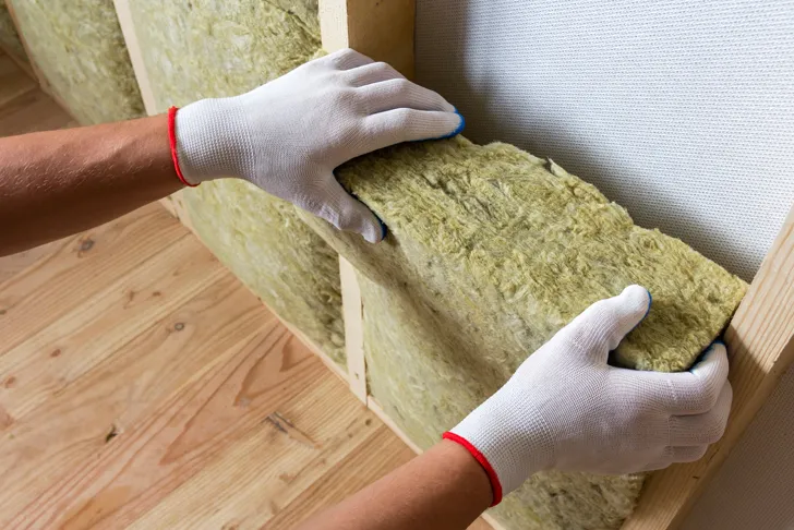 Ein Handwerker mit weißen Handschuhen installiert eine Wärmedämmung in Form von Mineralwolle zwischen Holzrahmen. Die Dämmung dient zur Verbesserung der Energieeffizienz und zur Reduzierung von Wärmeverlusten in Gebäuden.