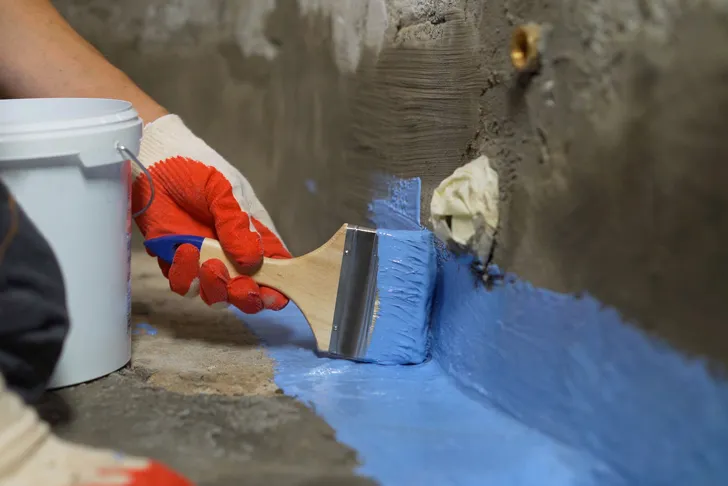 Bautenschutz durch das Auftragen einer blauen Abdichtungsmasse. Eine Hand mit Arbeitshandschuh führt einen breiten Pinsel entlang einer Wand. Die Beschichtung wir im unteren Wandbereich aufgetragen.