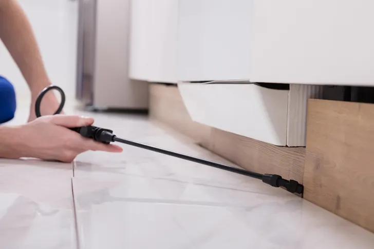 Schädlingsbekämpfer bringt Insektizid gegen Kakerlaken unter einem Küchenschrank auf Fliesenboden aus.