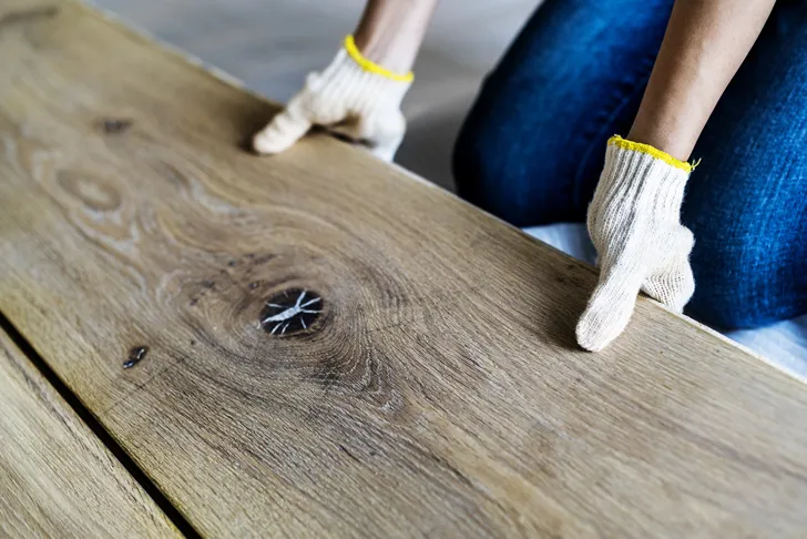 Ein Schreiner verlegt in einem Haus einen Holzboden und richtet eine massive Holzdiele mit Arbeitshandschuhen aus.