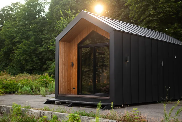 Modernes Tiny House mit schwarzer Metallfassade und Holzfront, großer Glasfront und Satteldach, gelegen am Waldrand im Abendlicht.