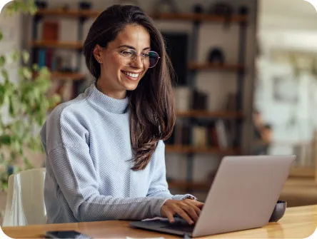 Junge Frau mit Brille und hellblauem Pullover sitzt für die Suche nach einem Gartenbauer in ihrer Nähe vor einem Laptop in einer hellen Wohnung